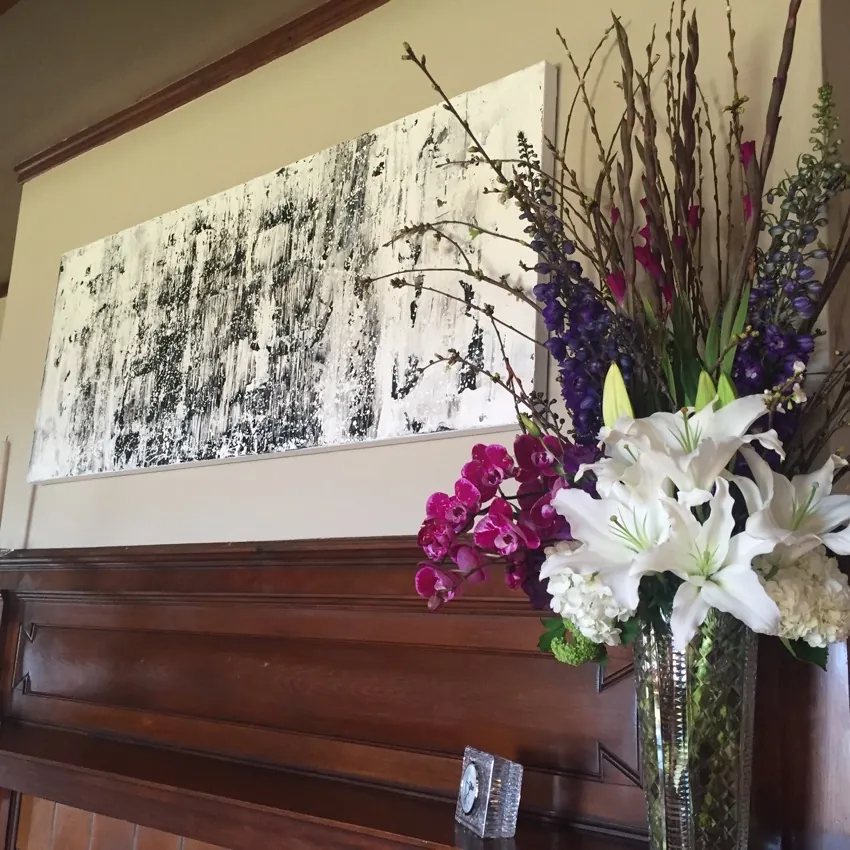 A modern abstract black-and-white painting, thoughtfully placed by an interior designer, hangs above a wooden mantel beside a tall glass vase filled with white lilies, purple and magenta flowers, and tall branches.