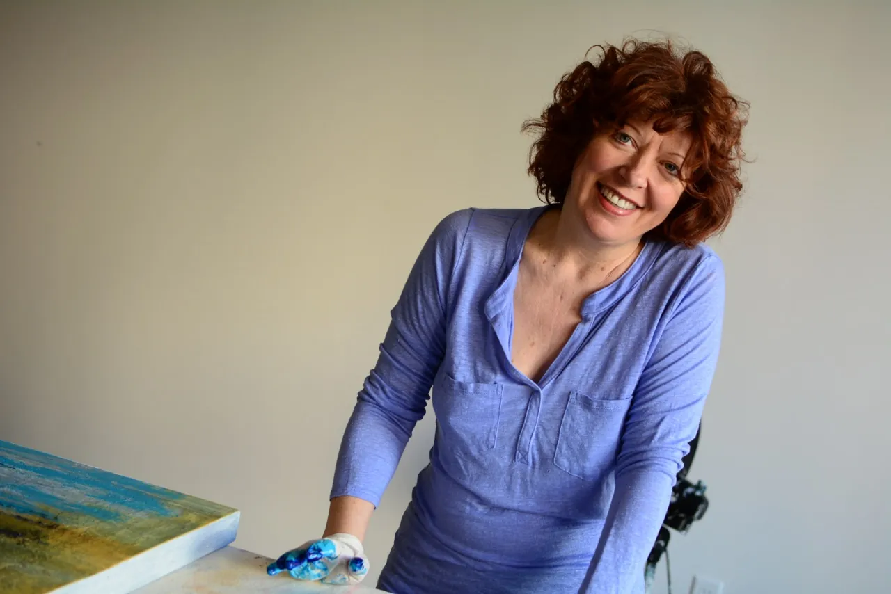 Abstract artist Clara Berta smiling in a blue shirt while painting a vibrant, large-scale original canvas in her California art studio.