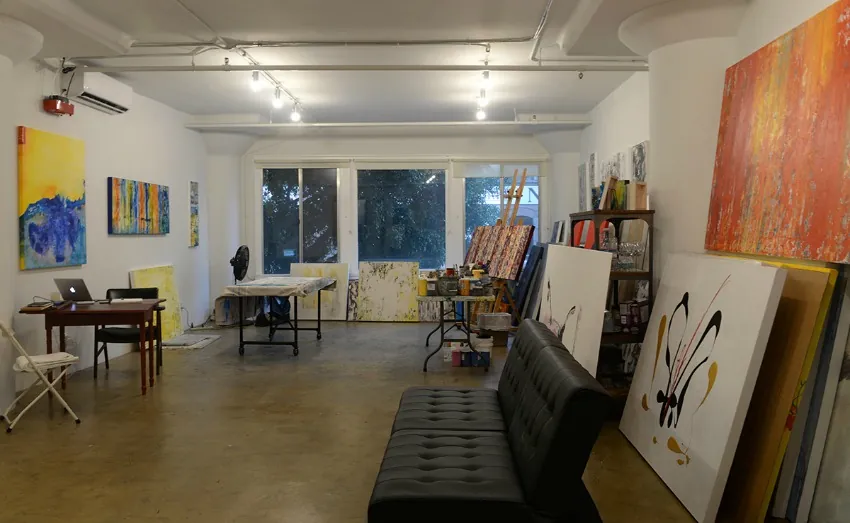 A spacious art studio with large windows, abstract paintings by contemporary abstract artist Clara Berta on the walls and floor, a black sofa, a table with art supplies, and a small desk with a laptop—perfect for an inspiring artist studio tour.