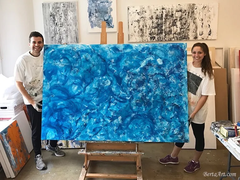 Two people in a bright Los Angeles painting class smile while holding a large abstract painting with vibrant blue and white swirls. Several other abstract artworks are displayed on the walls and floor around them.