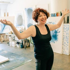 Artist Clara Berta stands with a warm, welcoming smile in the center of her bright, sun-filled Laguna Beach art studio. She is surrounded by her large-scale fluid acrylic paintings on white walls. The space is filled with natural light, creating an inviting atmosphere that connects her art with the serene studio environment.