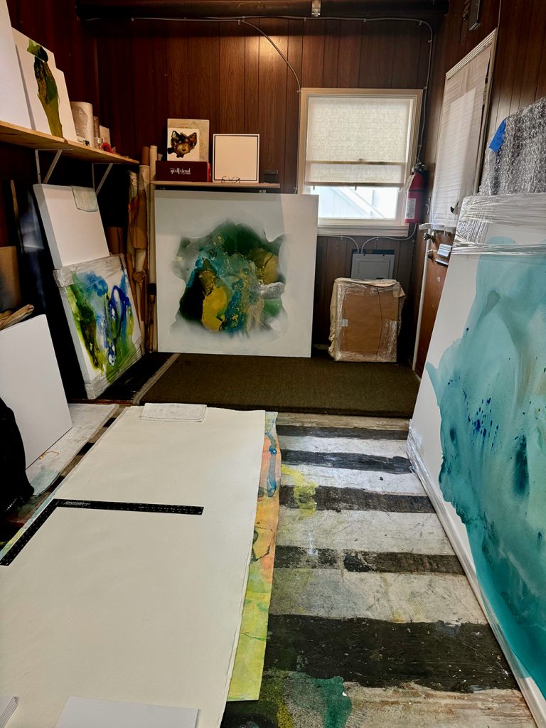 An art studio with wood-paneled walls, abstract paintings leaning against the walls, canvases, art supplies on shelves, and a large sheet of white paper on the floor with a ruler and colorful marks nearby.