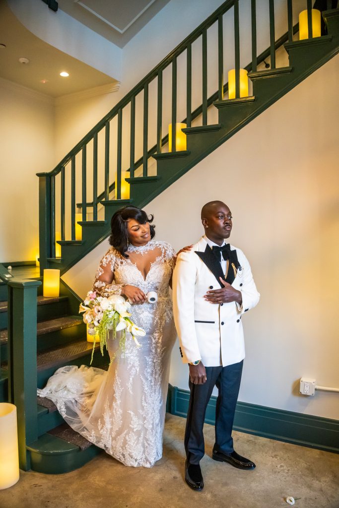 Bride and groom posing on staircase decorated with large glowing waxpillar candles, elegant flameless wax candle art rental for wedding venue decor