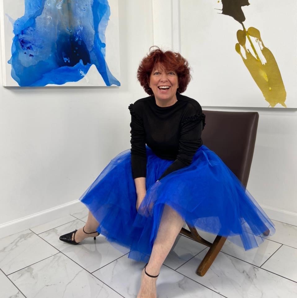 Artist Clara Berta smiling brightly while seated in a gallery, wearing a vibrant royal blue tulle puffy skirt and a black long-sleeved top. She is framed by two of her abstract paintings: a fluid blue-and-white piece on the left and a minimalist gold-and-black work on the right.