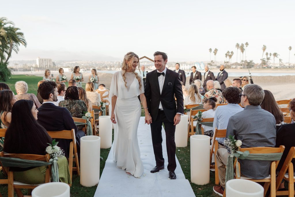 A bride and groom smile during their wedding recessional on a beachfront, walking down an aisle lined with oversized white paper luminaries glowing with warm light at dusk.