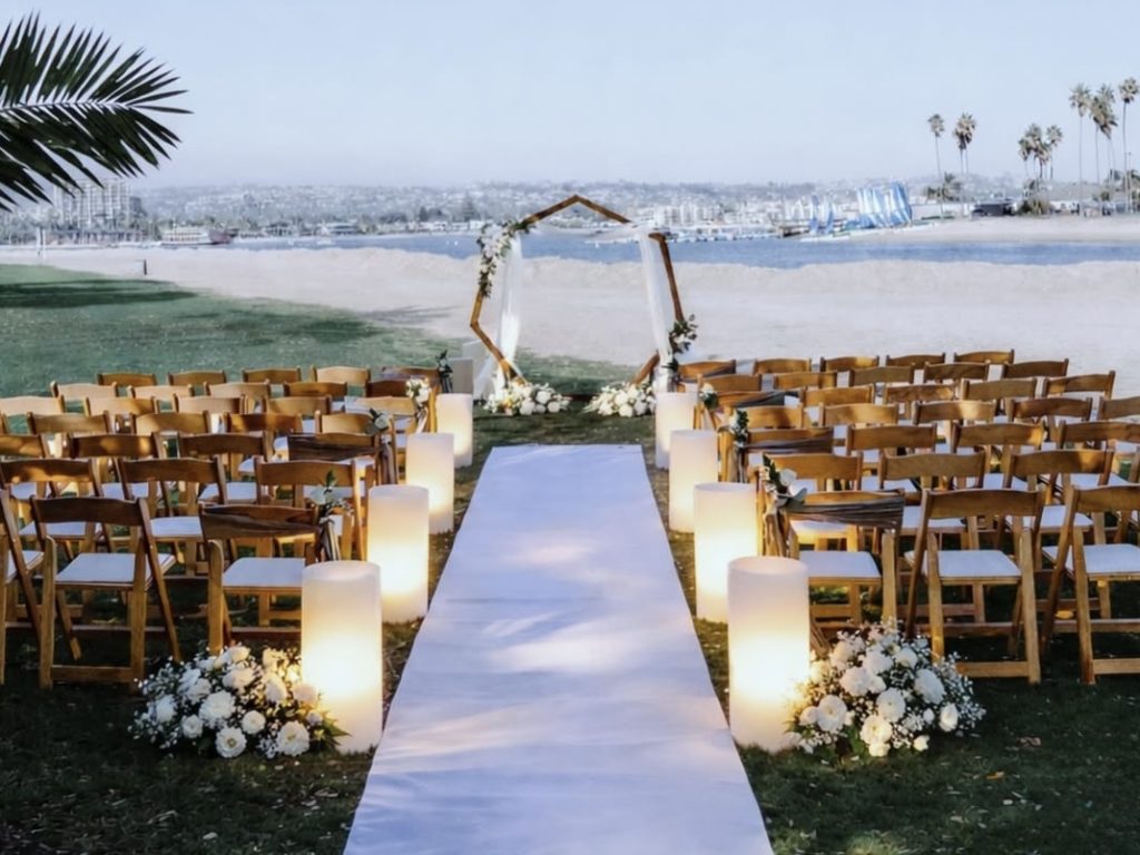Large, modern LED wax pillar candle sleeves lining a white wedding aisle. These oversized, battery-operated wax cylinders glow warmly from within, alternating with white floral bouquets on a grassy lawn leading to a coastal ceremony.