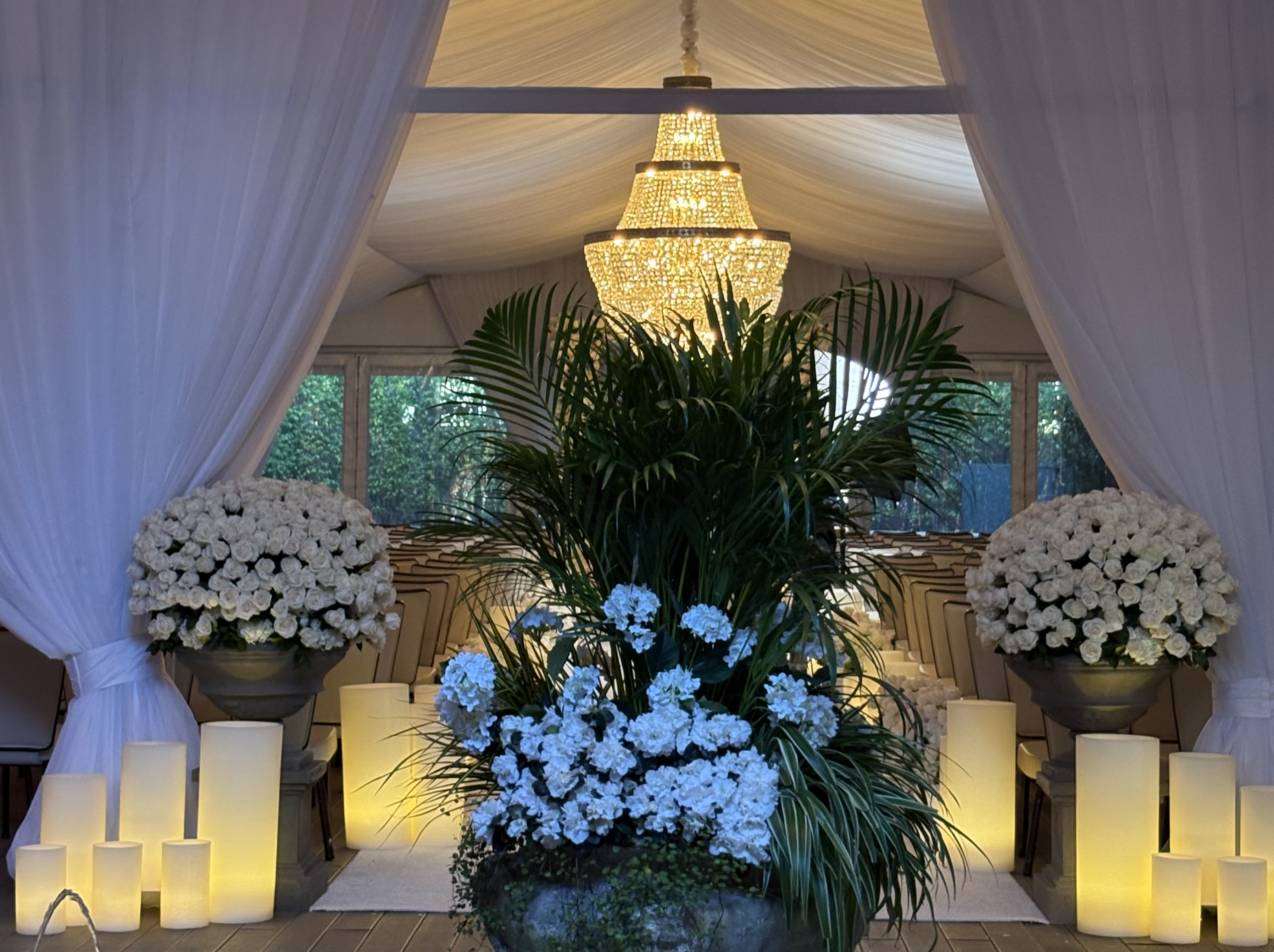 The wedding ceremony aisle was illuminated with large abstract wax flamelesscandle art arrangements and white floral decor under crystal chandelier in a draped tent venue