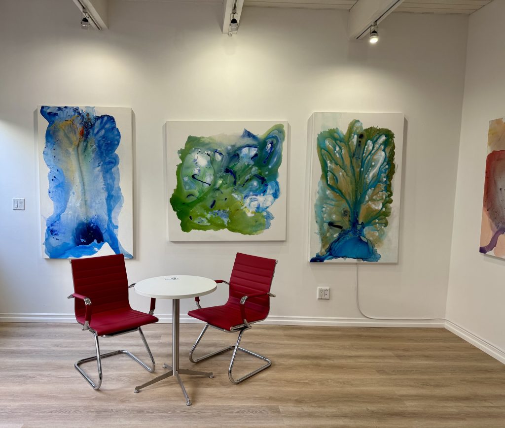 Modern gallery interior of Clara Berta Studio Laguna Beach with three blue and green abstract paintings on a white wall above two red chairs.
