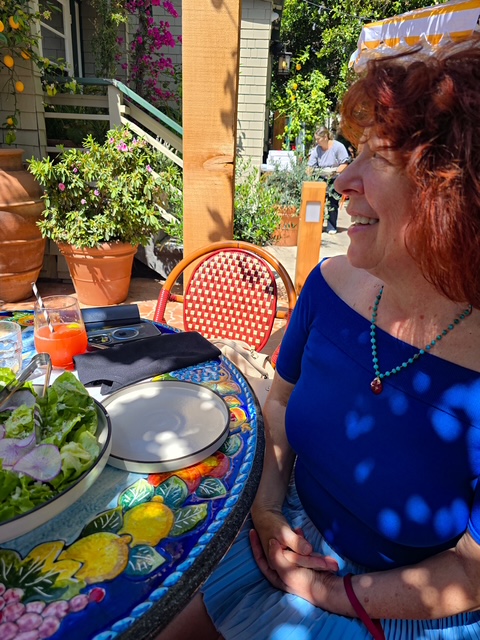 Artist Clara Berta dining at Truly Pizza Laguna Beach; a bright, sunlit portrait featuring a blue blouse, artisan pizza on a silver tray, and a beautiful Mediterranean-style setting with lemon trees.