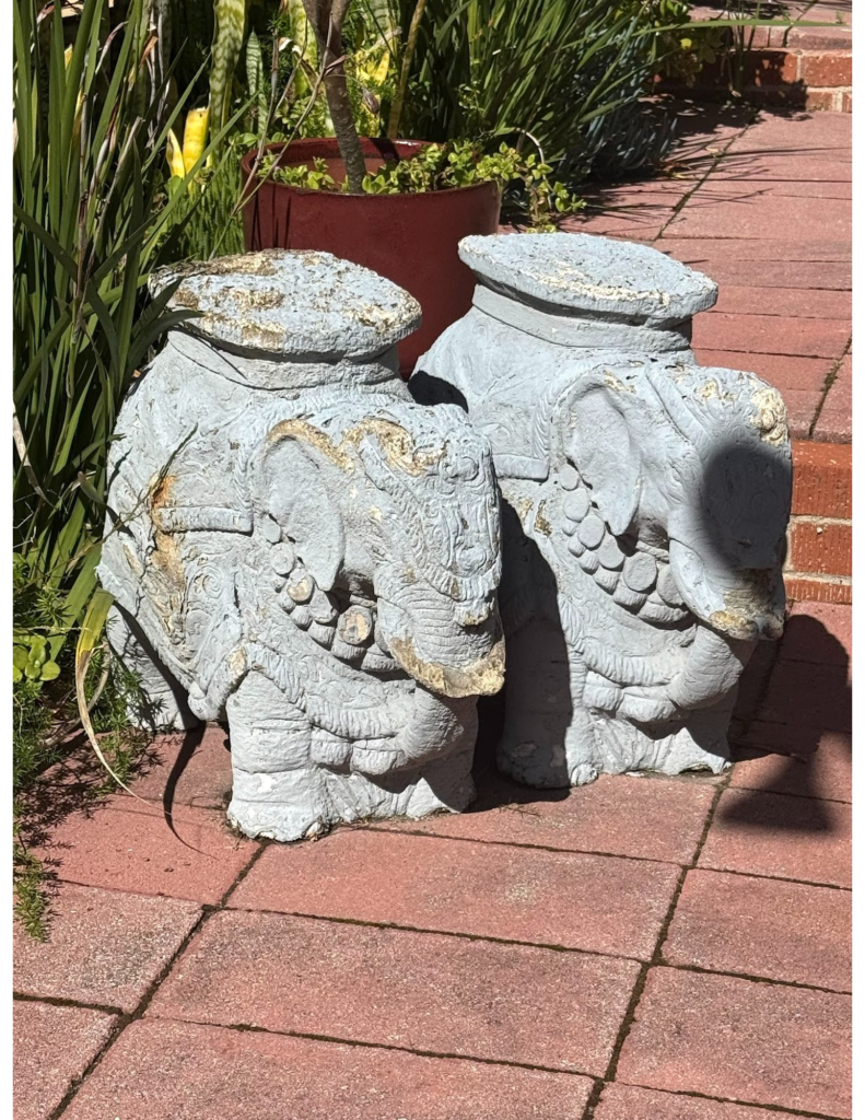 Two vintage elephant sculptures with an aged patina standing in the lush garden of a Laguna Beach art studio, surrounded by green foliage.