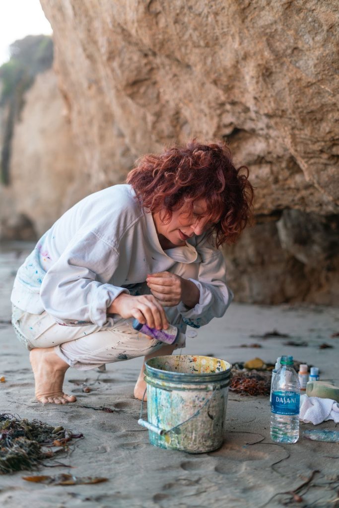 Artist Clara Berta immersed in her creative process at the beach, working with a bucket of paint against the backdrop of ocean waves.