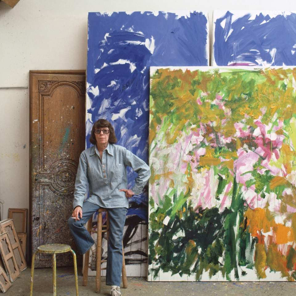 Legendary abstract expressionist artist Joan Mitchell standing in her studio, framed by her monumental, multi-panel paintings featuring wild, gestural brushstrokes of vibrant blues and yellows.