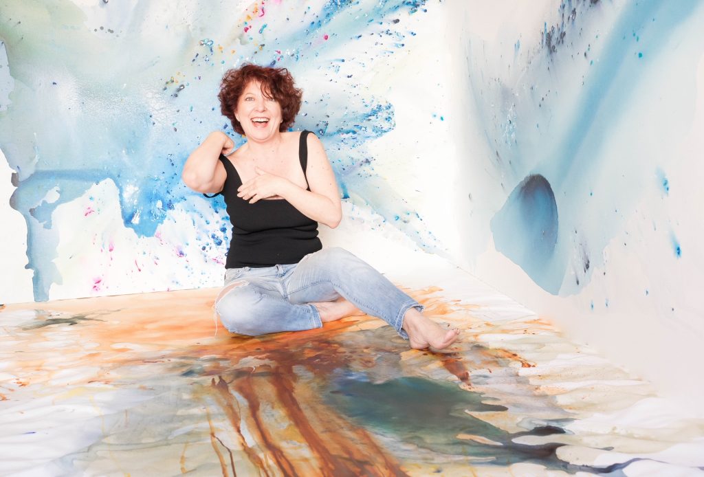 Artist Clara Berta, smiling and barefoot, sits surrounded by her calming abstract paintings within an immersive, custom-painted studio corner featuring blue, gold, and green fluid acrylic washes and airbrush effects.