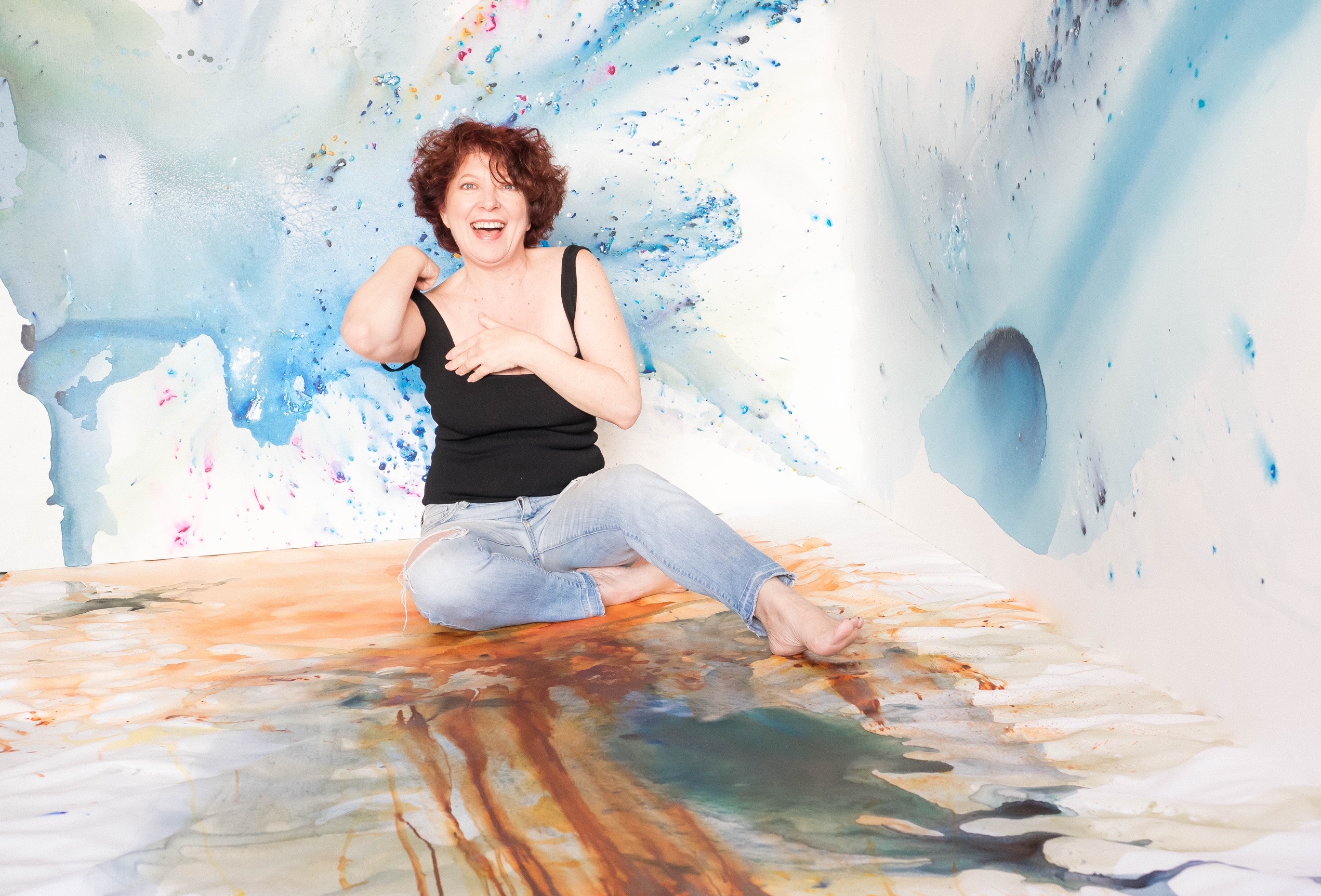 Artist Clara Berta, smiling and barefoot, sits surrounded by her calming abstract paintings within an immersive, custom-painted studio corner featuring blue, gold, and green fluid acrylic washes and airbrush effects.