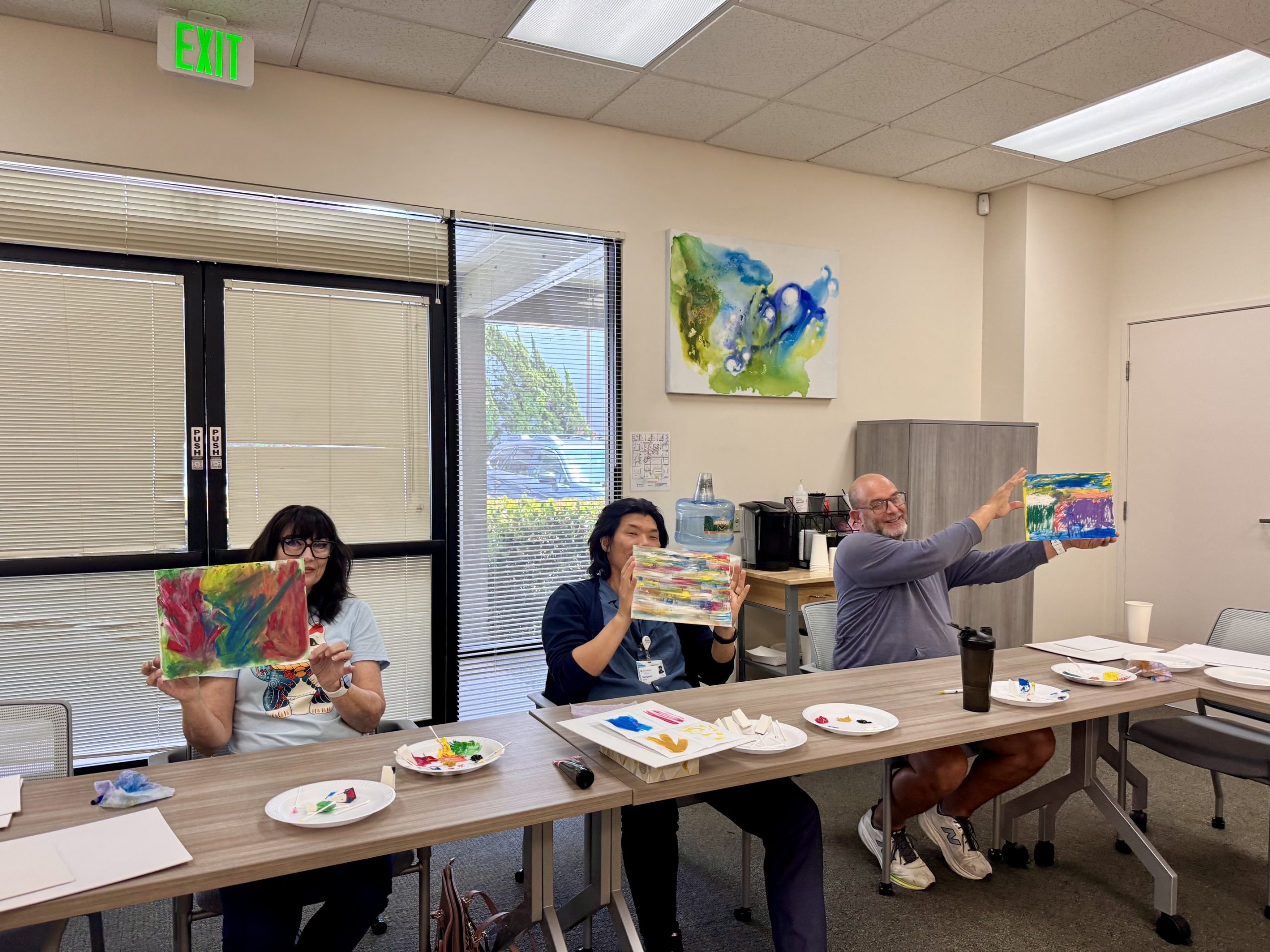 A diverse group of happy students sitting at tables in an Art4Healing classroom, smiling and holding up their unique, vibrant acrylic paintings. The artwork features expressive, colorful abstract designs, reflecting a positive and therapeutic creative session.