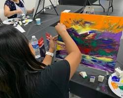 A tight close-up of a student's hand using a wedge-shaped makeup sponge to smoothly blend soft layers of pink and blue paint onto a canvas during an Art4Healing class.