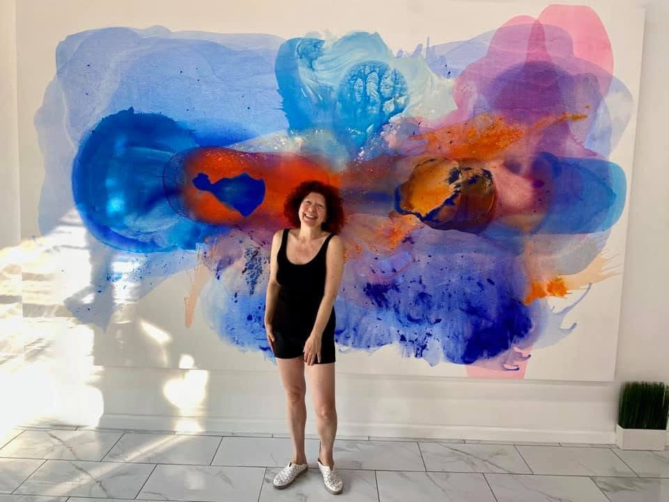A monumental abstract fluid painting titled 'Celebration' measuring 72x115 inches by artist Clara Berta. The vibrant, large-scale composition features an explosive burst of translucent magenta, deep cobalt blue, brilliant yellow, and soft violet acrylic washes that dynamically splash and bleed outward against a stark, minimalist white background.