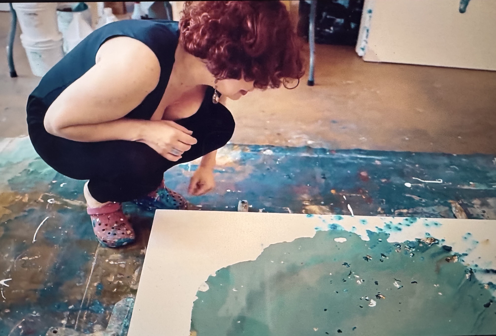 Artist Clara Berta in her studio, demonstrating her signature action-painting process. A large abstract canvas rests flat on the floor as she carefully pours vibrant, fluid acrylic paint from a container, letting the colors swirl and bleed together naturally across the surface.
