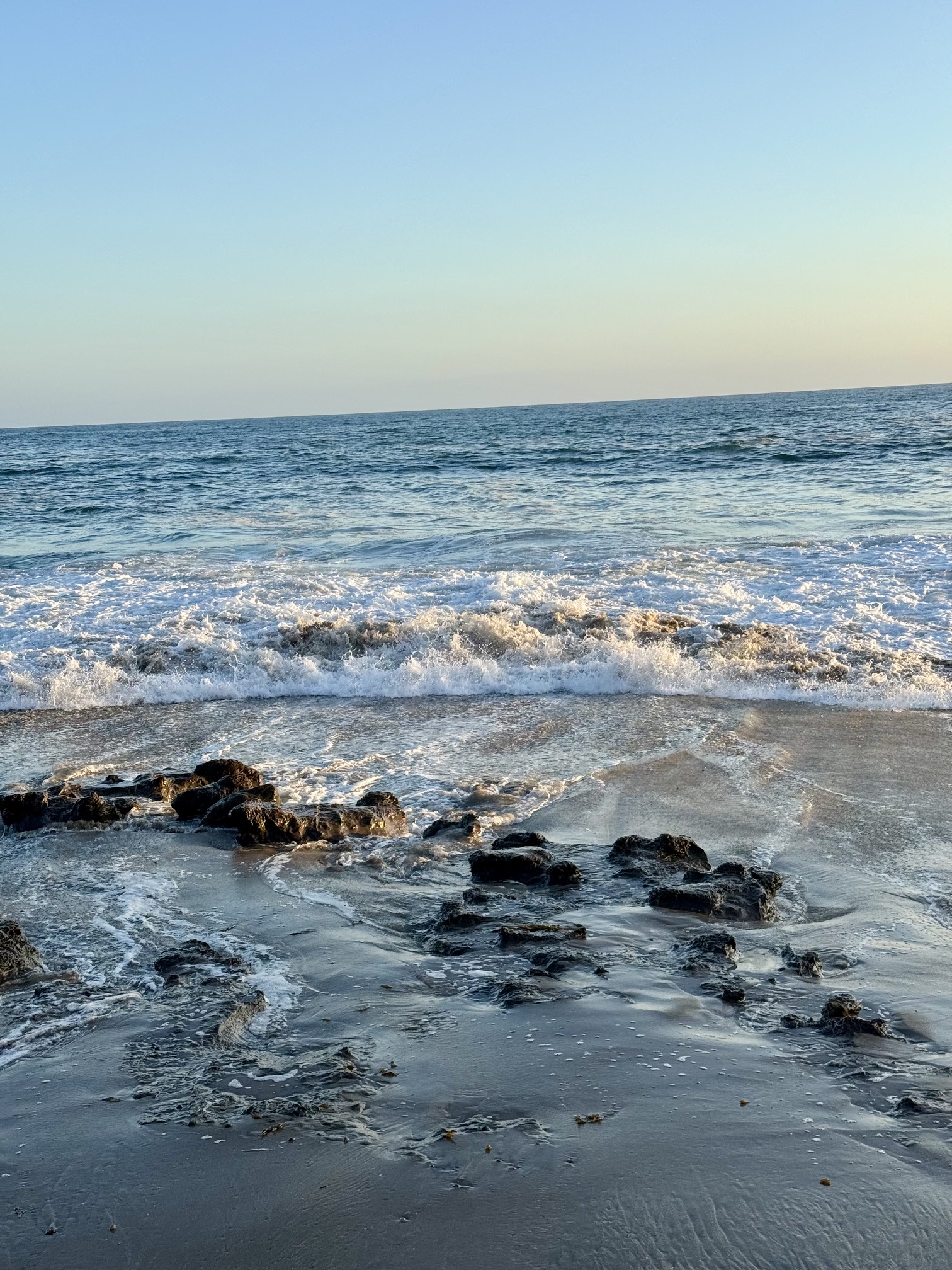 A wide shot of a coastal beach at high tide, with powerful ocean waves crashing against a sandy shoreline under a clear blue sky.