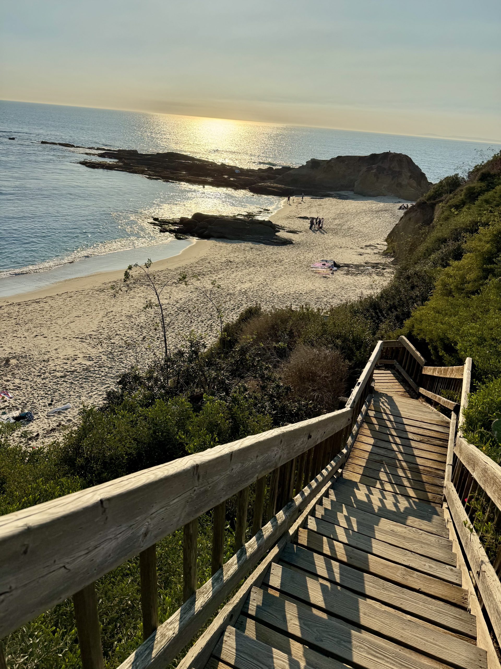 A wooden staircase leading down a steep cliffside to a sandy beach in Laguna Beach, California, with the blue ocean waves crashing in the background.