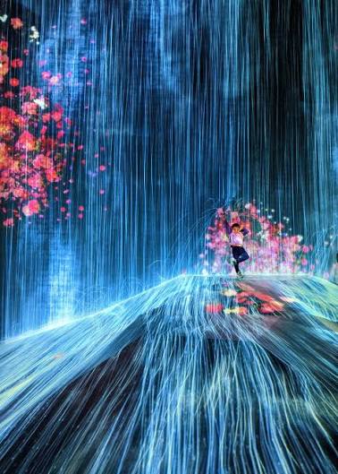 A person stands atop a curved hill inside a digital art exhibit at teamLab Planets Tokyo, surrounded by vertical, glowing blue light projections resembling a waterfall with pink floral accents.