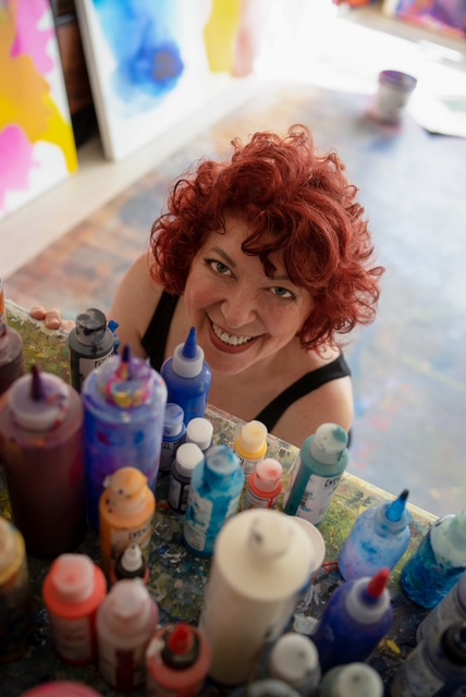 Abstract artist Clara Berta in her studio, surrounded by a vibrant collection of professional-grade acrylic paints, preparing for her next large-scale canvas.