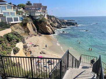 An overlook view of Woods Cove in Laguna Beach, featuring stone steps leading down to a secluded sandy cove with turquoise ocean waves rolling onto the shore, framed by rugged coastal rocks.
