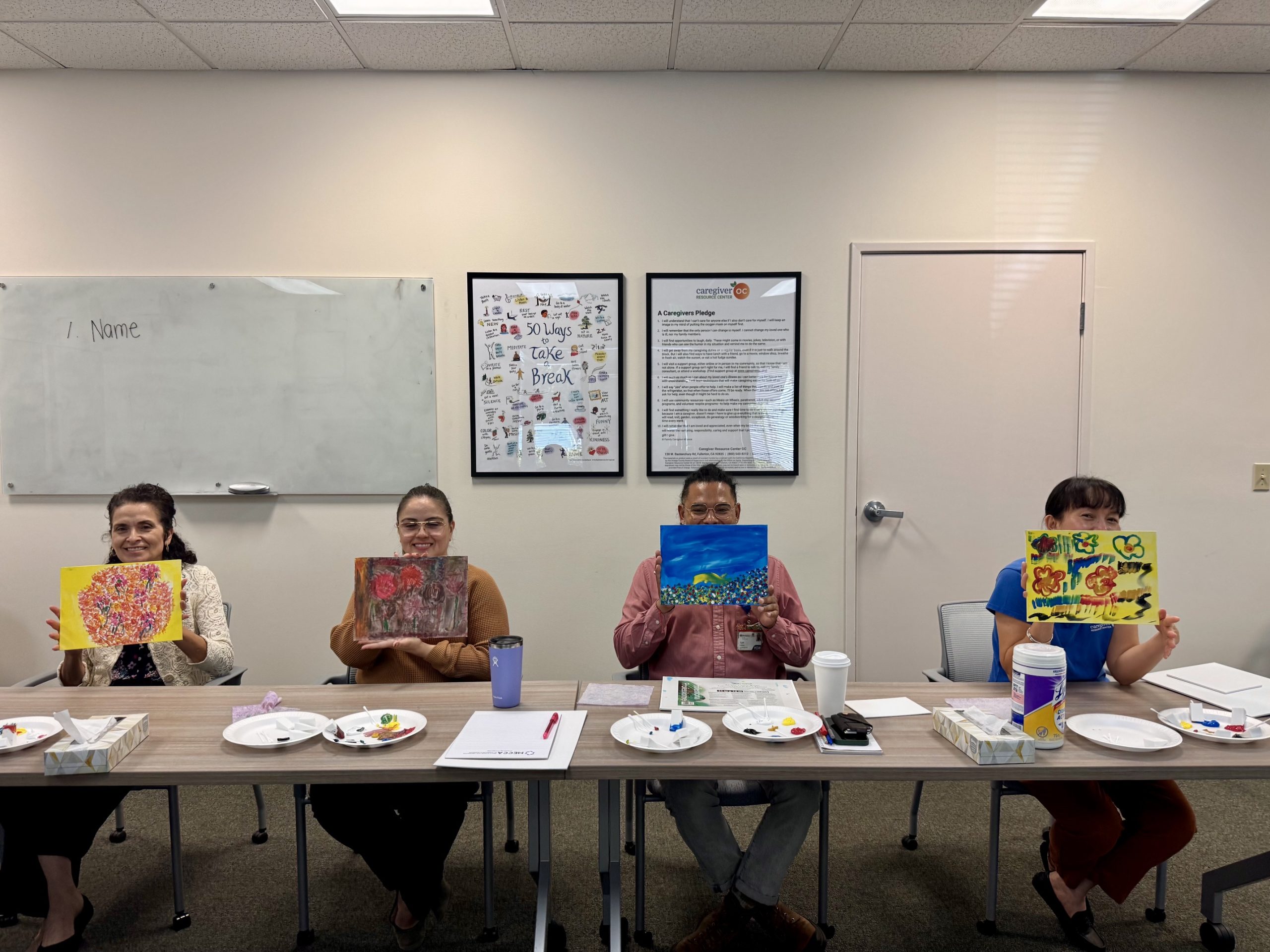 A row of seated Art4Healing participants holding up their finished canvases. Each painting displays a completely unique mix of colors and textures, reflecting each student's personal creative expression.