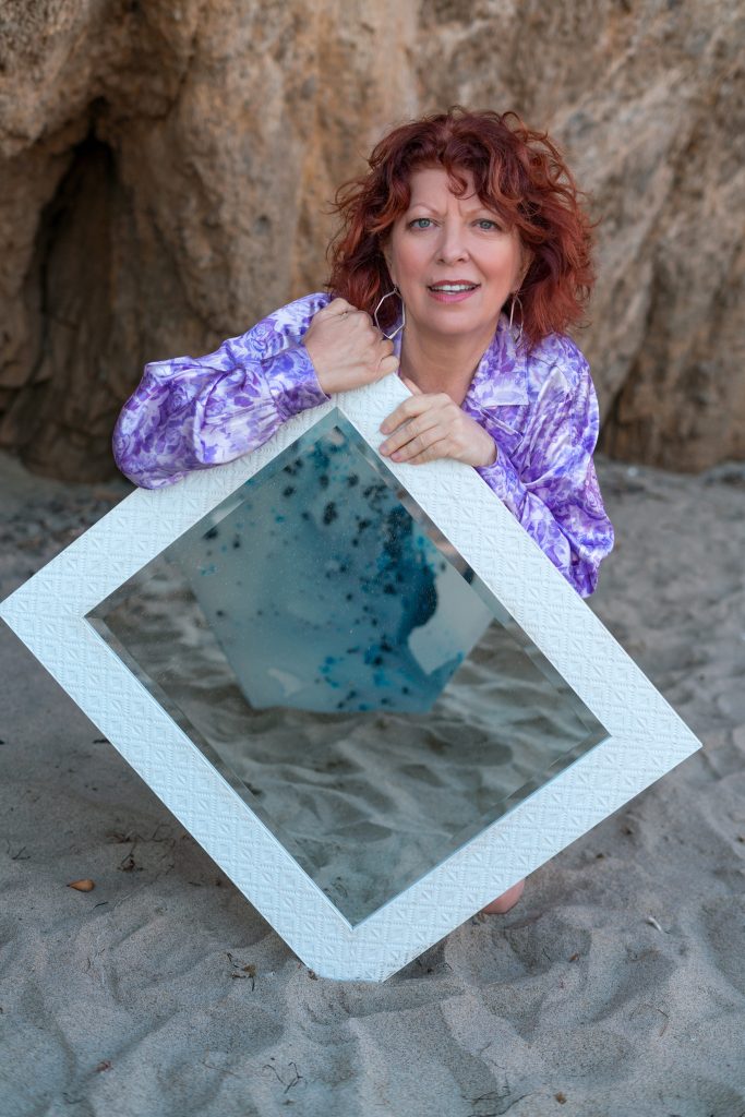 Abstract artist Clara Berta kneeling barefoot on the sand at El Matador Beach next to her modern abstract painting, featuring fluid seafoam green and ocean blue washes contrasting with textured, raised coral-pink details.