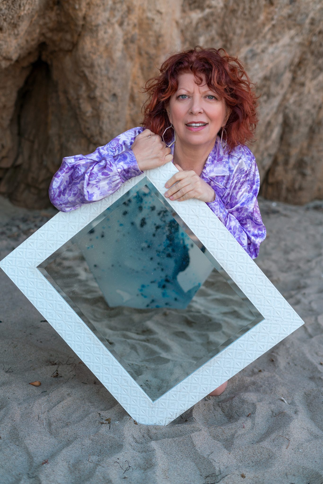 Abstract artist Clara Berta kneeling barefoot on the sand at El Matador Beach next to her modern abstract painting, featuring fluid seafoam green and ocean blue washes contrasting with textured, raised coral-pink details.