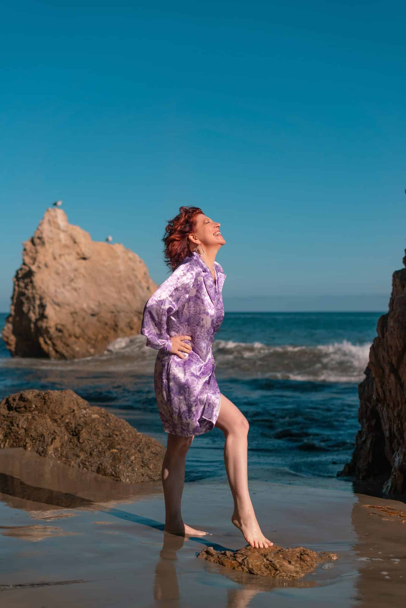 Clara Berta standing on the rugged, sandy shore at El Matador Beach in Malibu, surrounded by towering sea stacks and dramatic rock formations under a soft coastal sky.