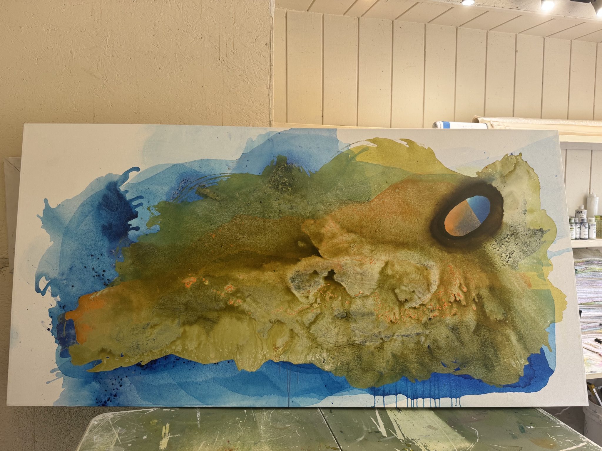 A fluid abstract painting in progress by Clara Berta, featuring organic washes of cerulean blue, olive green, and ocher yellow, with a distinct glowing peach and blue oval portal on the right.