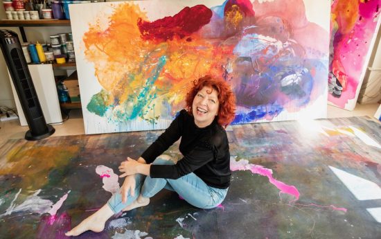 A smiling woman with curly red hair sits barefoot on a colorful, paint-splattered floor in front of a large, vibrant abstract painting in an art studio. She is wearing a black top and light blue jeans.