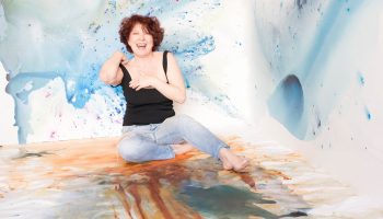 Artist Clara Berta, smiling and barefoot, sits surrounded by her calming abstract paintings within an immersive, custom-painted studio corner featuring blue, gold, and green fluid acrylic washes and airbrush effects.